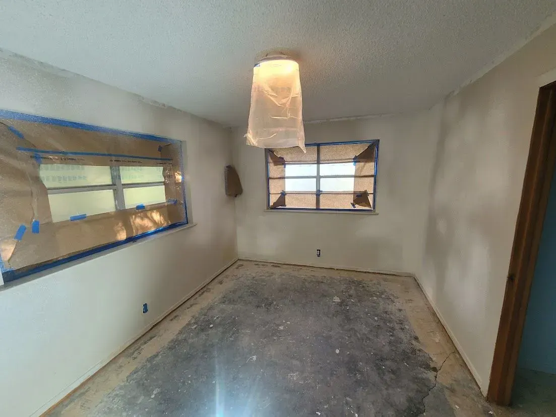 Empty room under renovation with bare floor, white walls, and two windows.