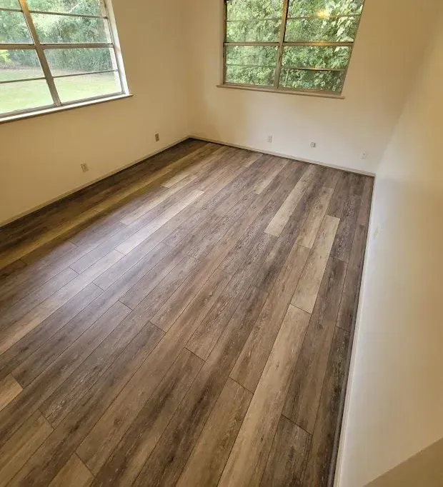 Empty room with wood-look flooring, cream walls, and two windows with trees outside.