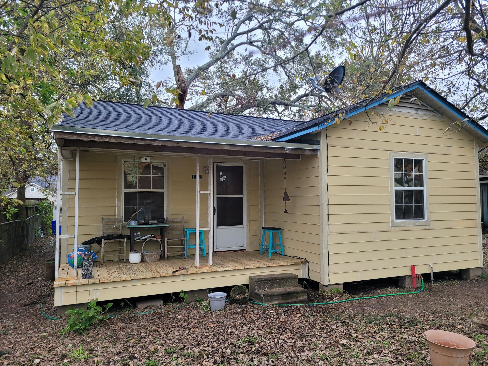 Small beige house with a blue roof, front porch, and two turquoise chairs under trees.