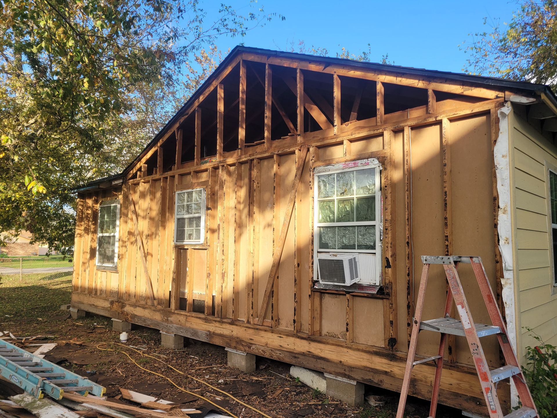 Partially renovated tan house with exposed framing and a ladder leaning against it outdoors