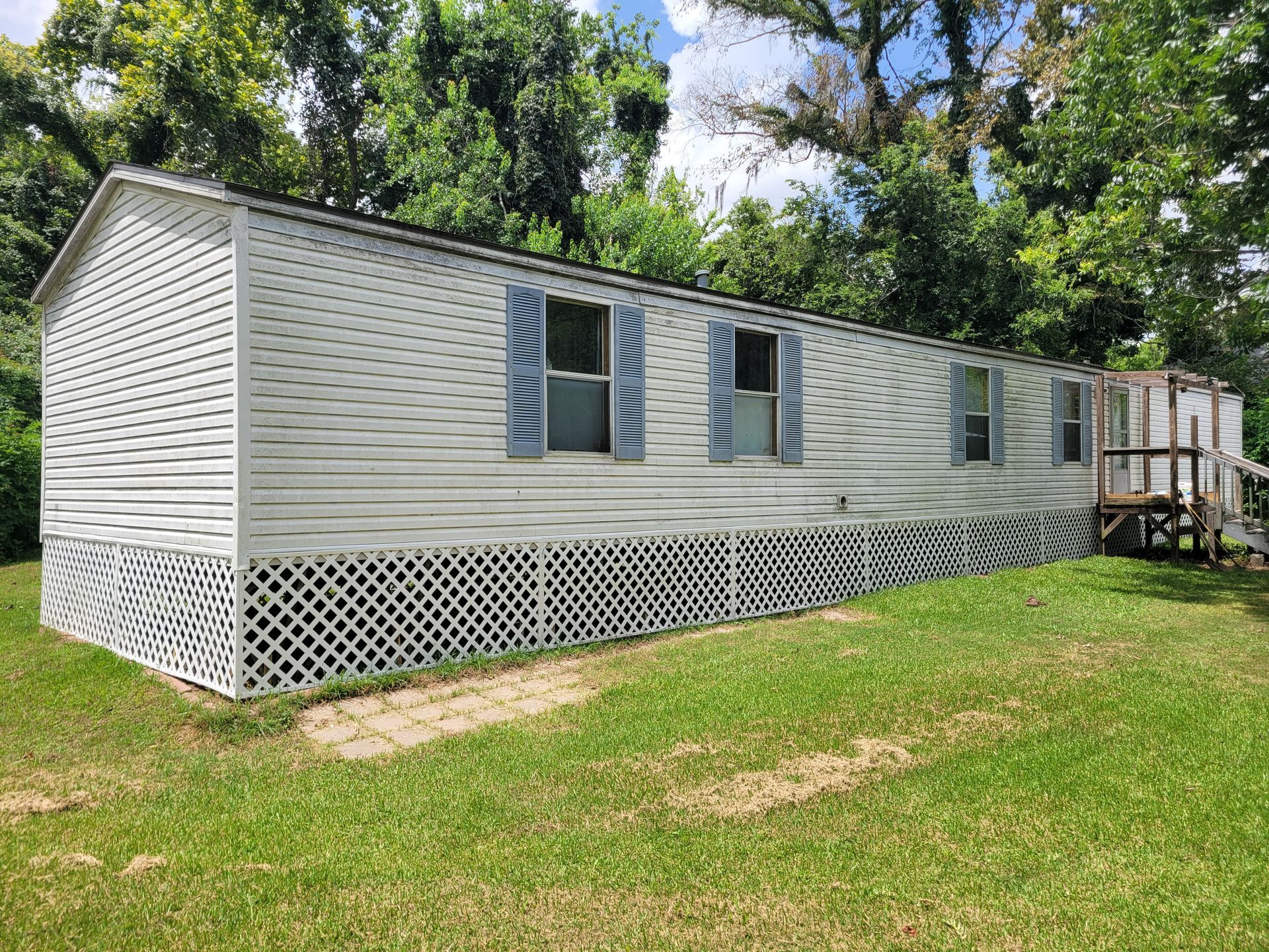 Gray mobile home with white lattice skirting, blue shutters, and a small porch in a grassy yard.