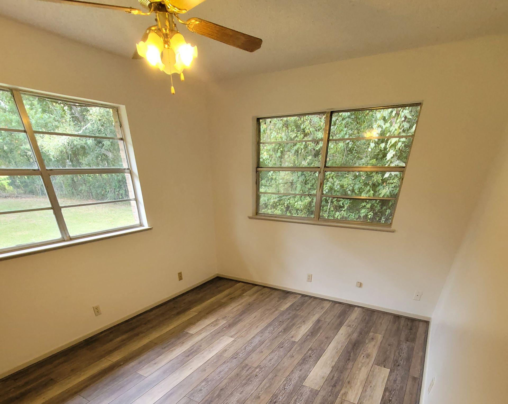Empty beige bedroom with wood-look flooring, two windows, and a ceiling fan light.