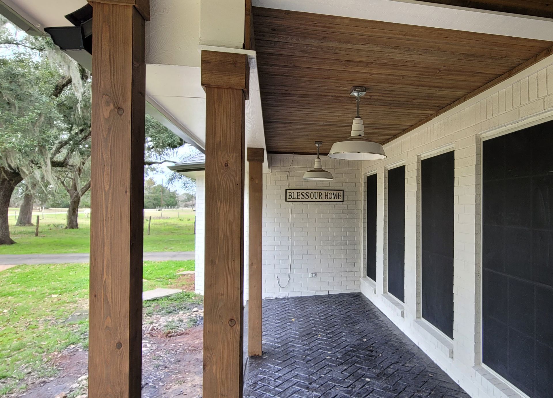 Covered porch with wooden post, hanging light, screened windows, and brick floor beside a lawn