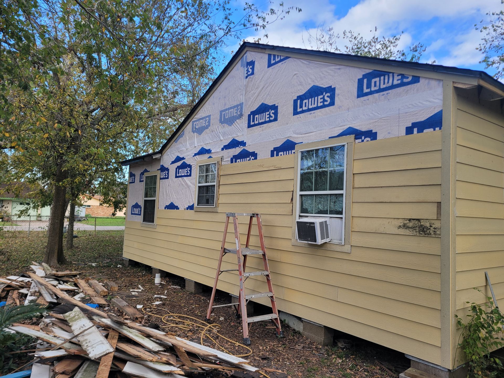 House exterior under renovation with beige siding, roof paper, a ladder, and debris on the ground