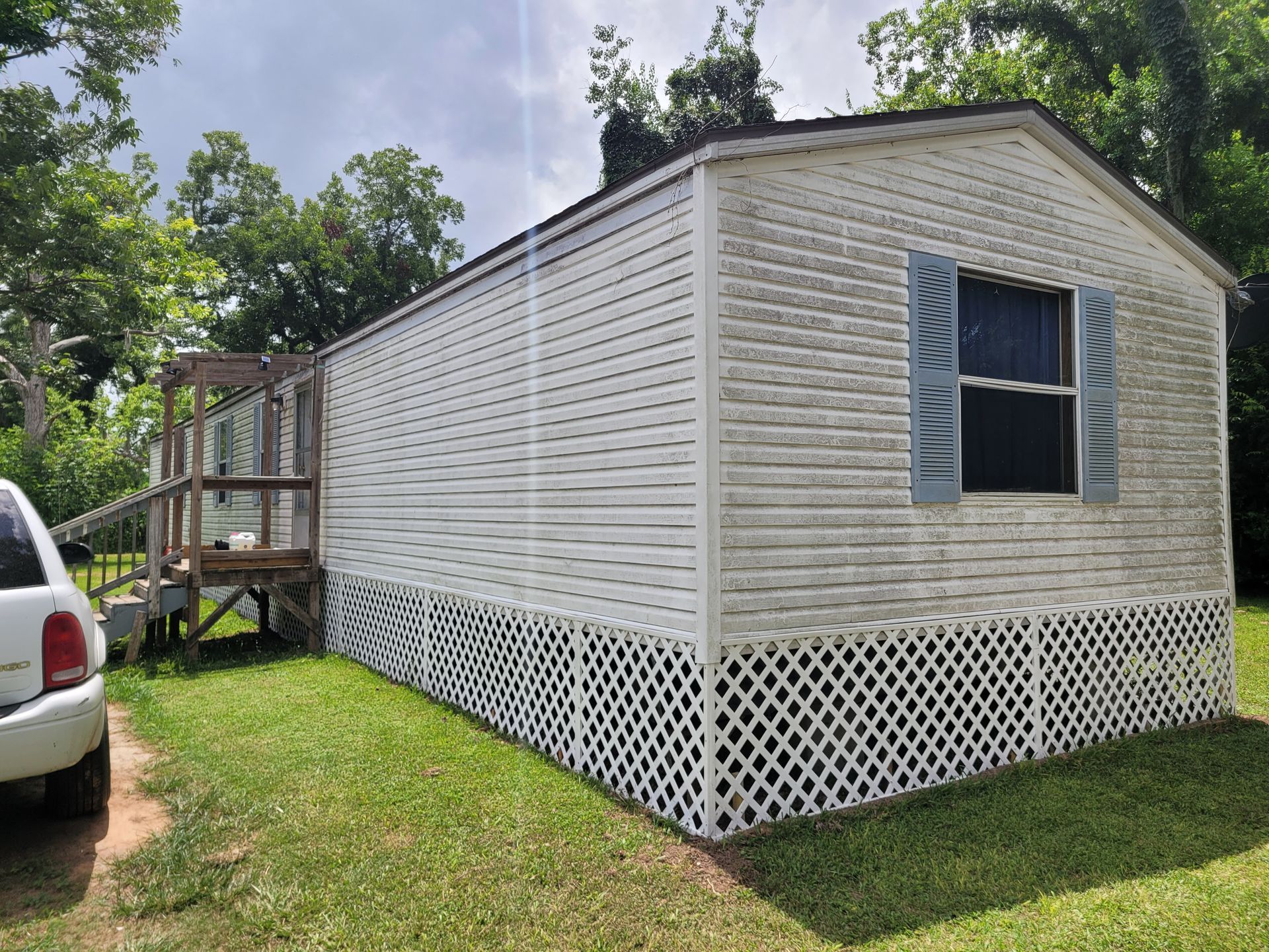 White mobile home with lattice skirting and a front window, set on a grassy lot beside a vehicle.