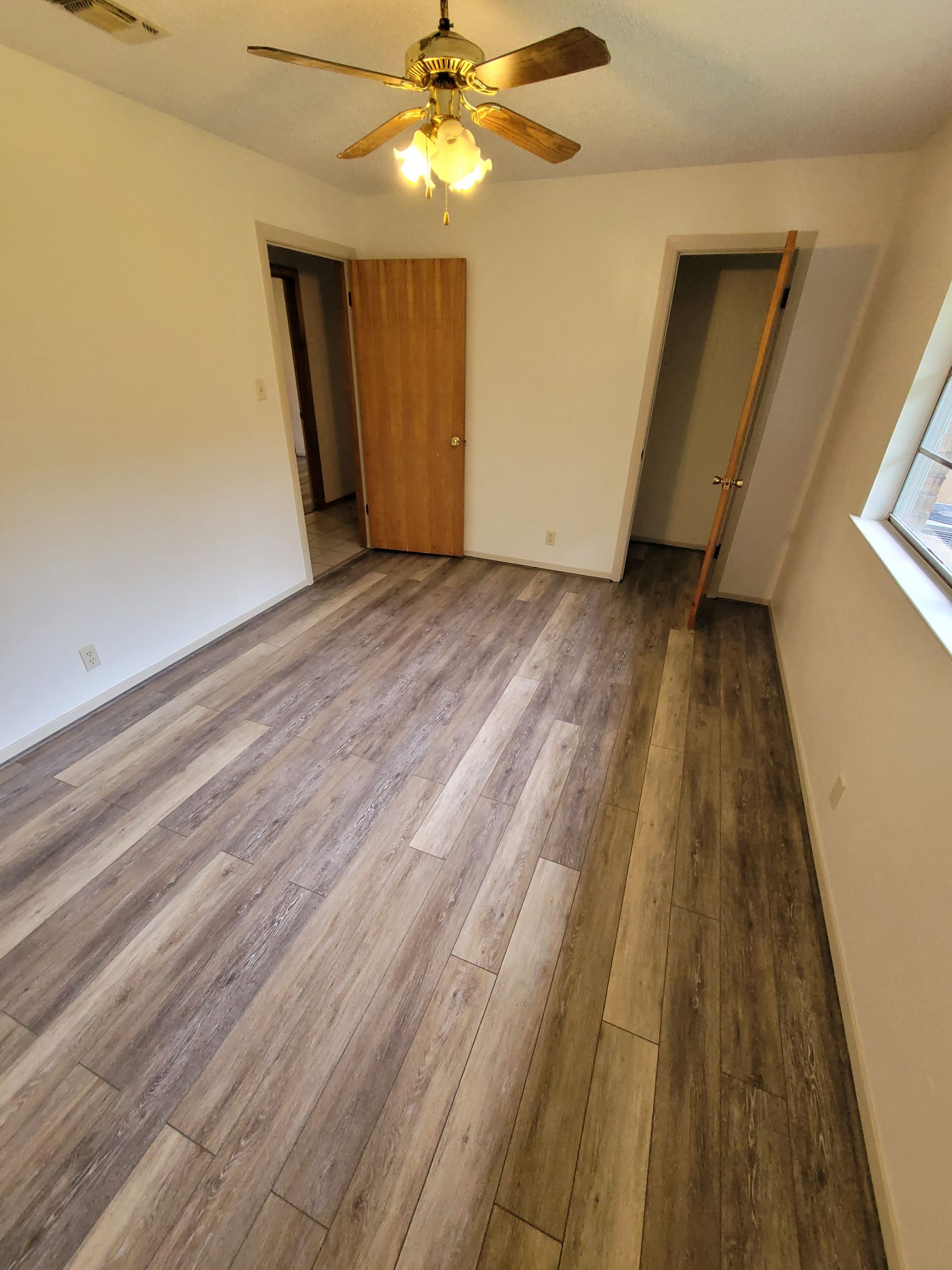 Empty room with wood flooring, ceiling fan lights, white walls, and a window on the right.