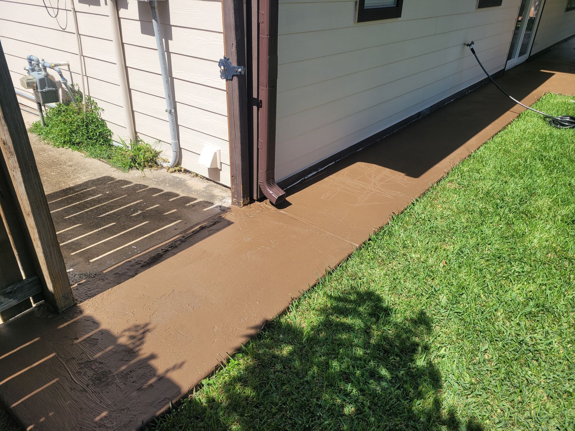 Backyard walkway beside a house, with a brown paved path and green grass in sunlight.