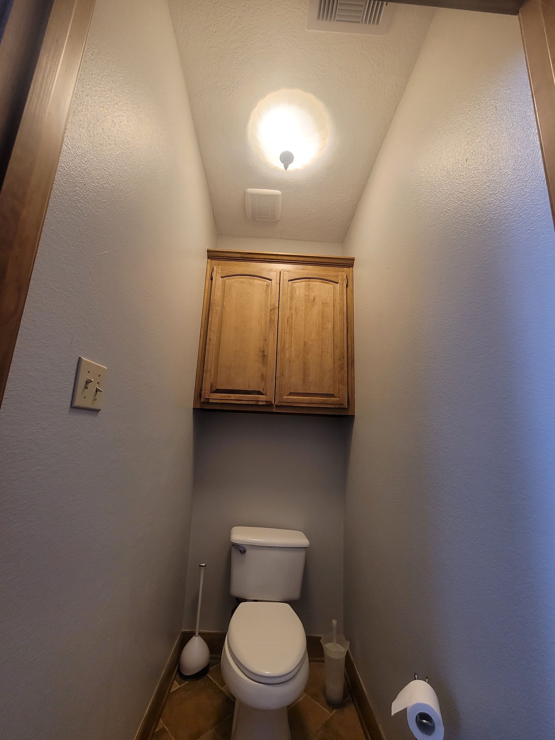 Narrow bathroom with toilet, wood cabinet above, and a single ceiling light.