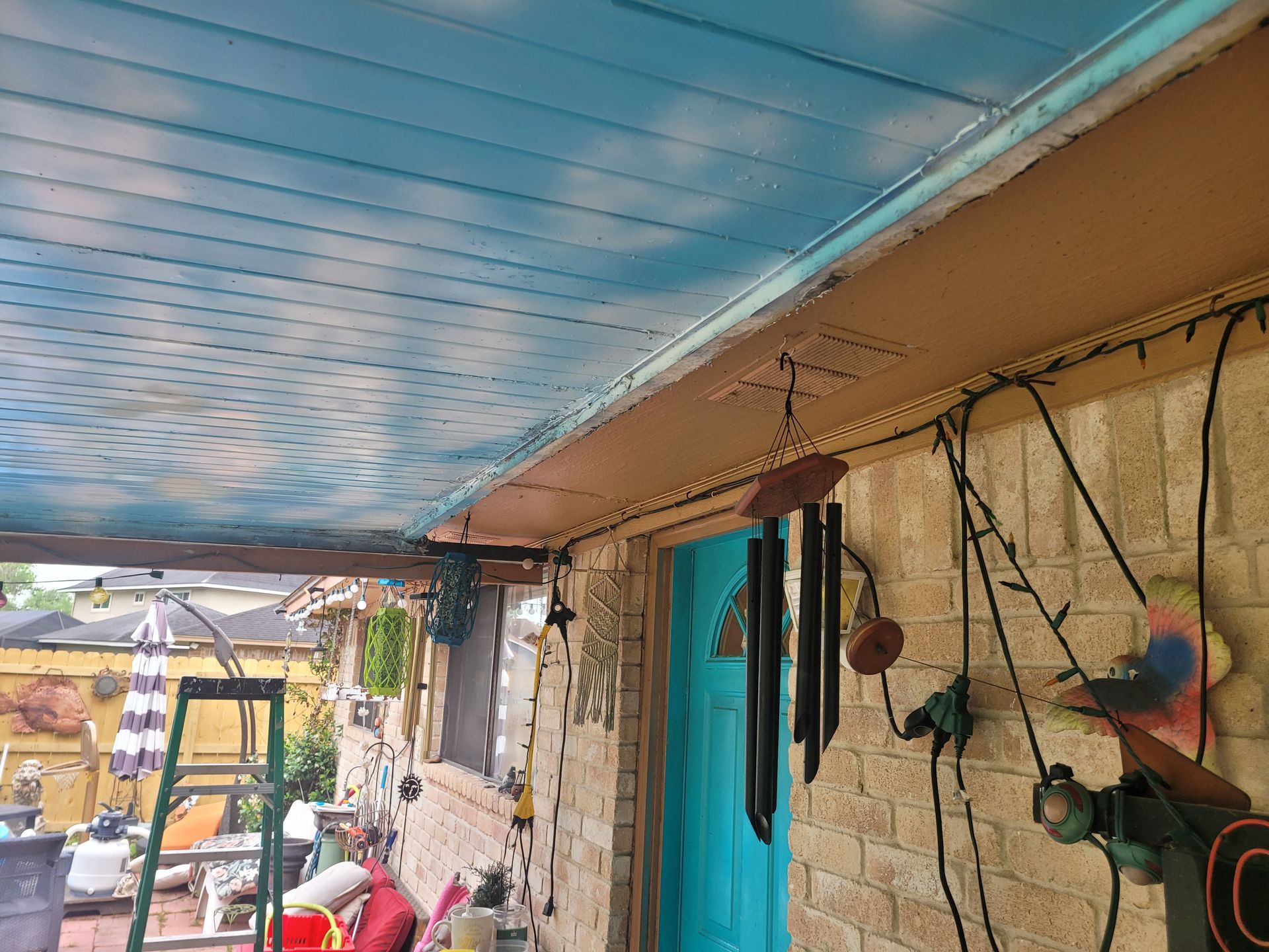 Narrow outdoor walkway with blue tarp roof, hanging cables, turquoise door, and tools on a rustic wall.