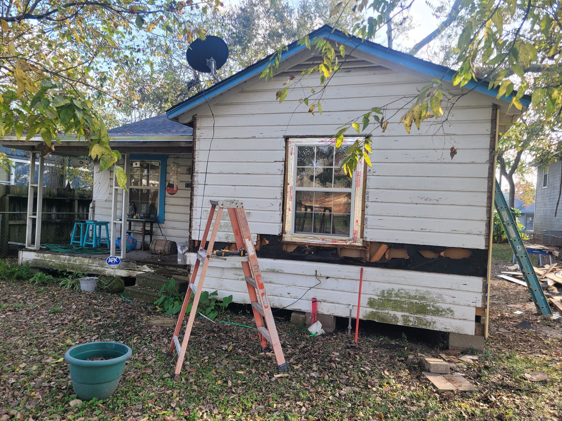 Small white house with peeling paint, ladder, and tools during exterior repair work in a leafy yard
