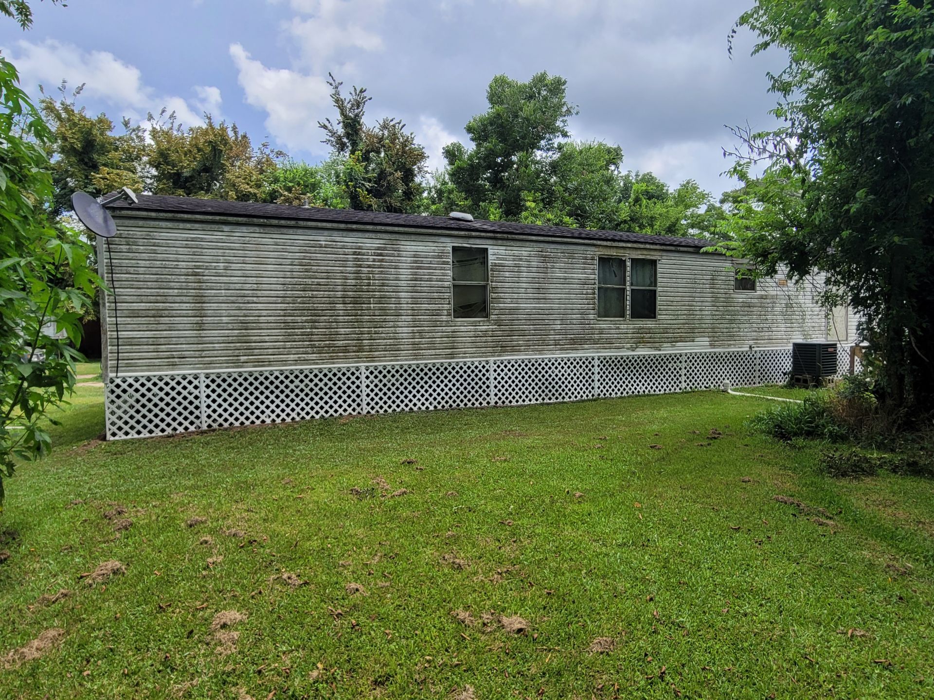 White mobile home with small windows and lattice skirting on a grassy lot under cloudy skies