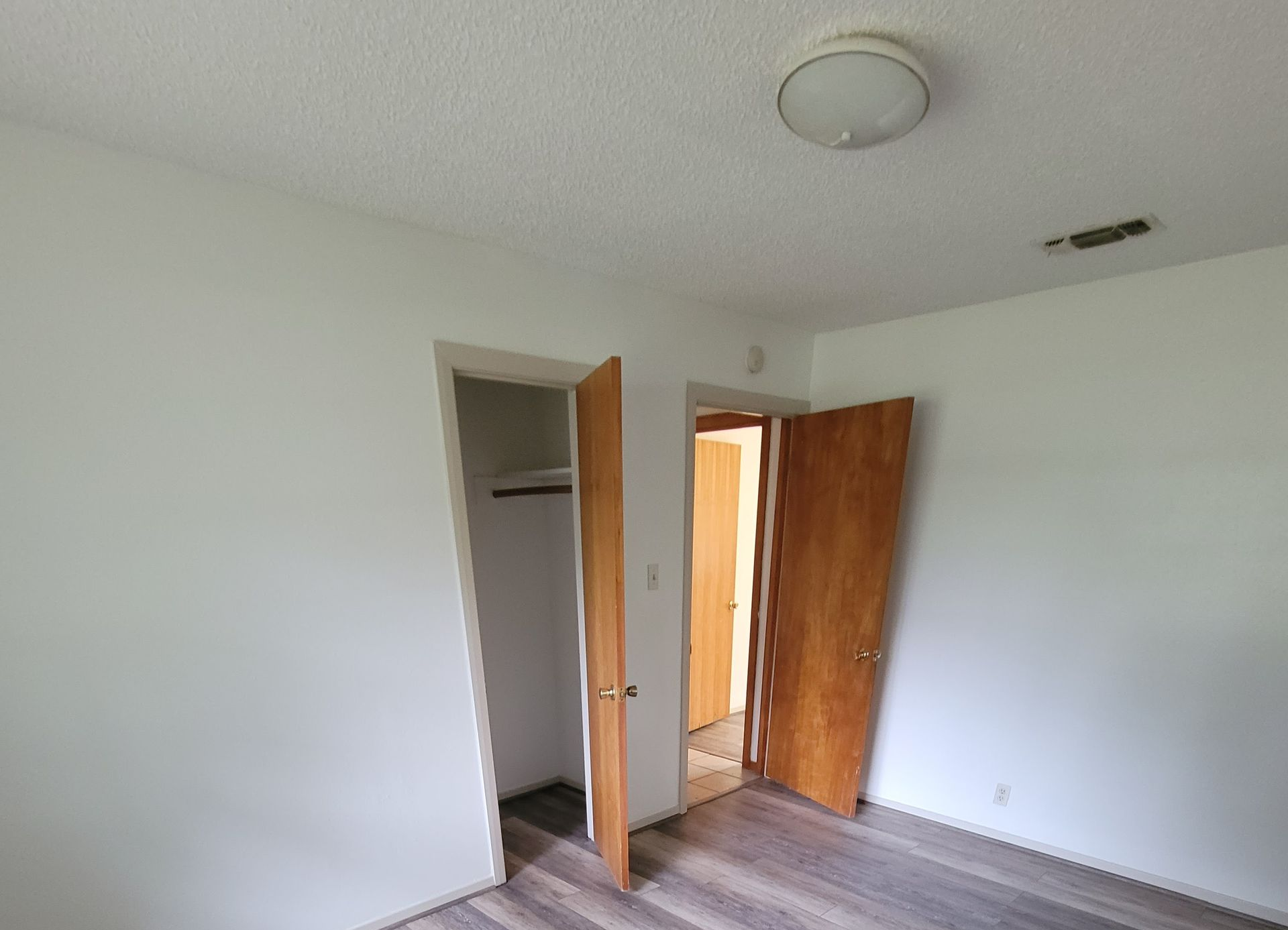 Empty white bedroom with open closet and door, wood flooring, and ceiling light fixture