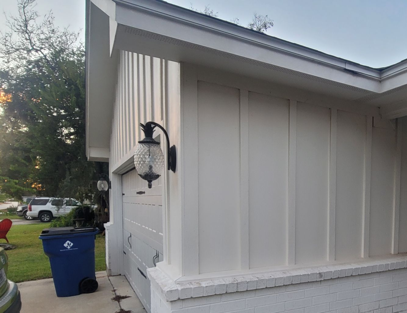 White house exterior corner with black lantern, blue trash bin by sidewalk, and a car in the background