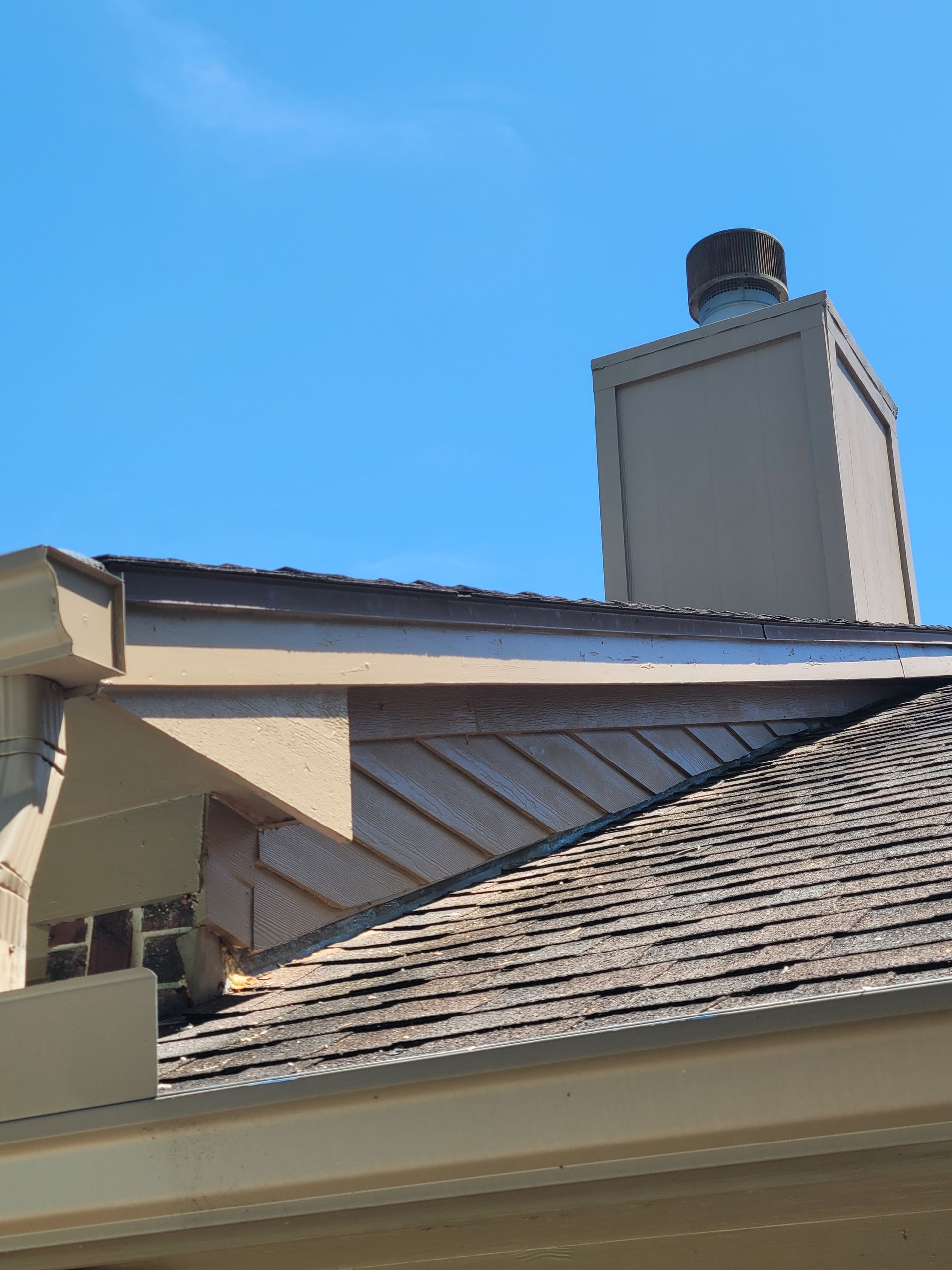 House roof with tan shingles and a white chimney against a clear blue sky