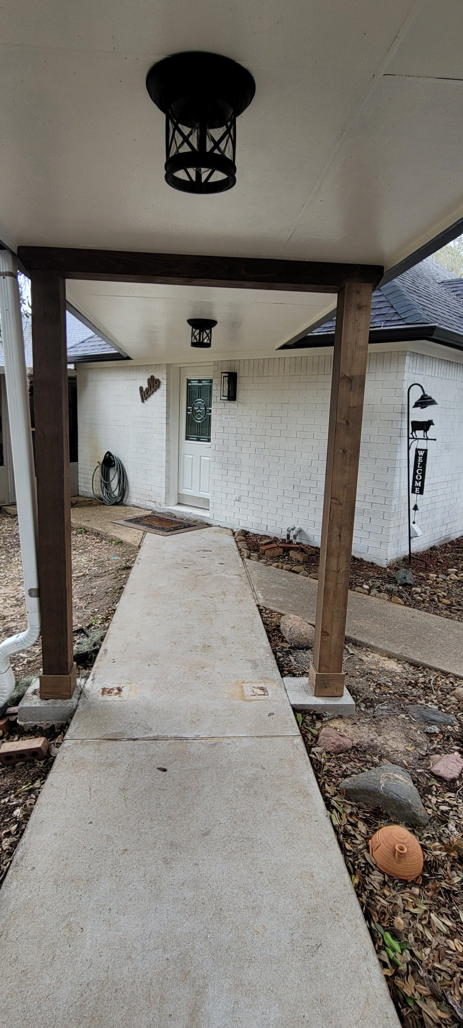 Covered concrete walkway leading to a white house entrance, with rustic wooden posts and black lanterns