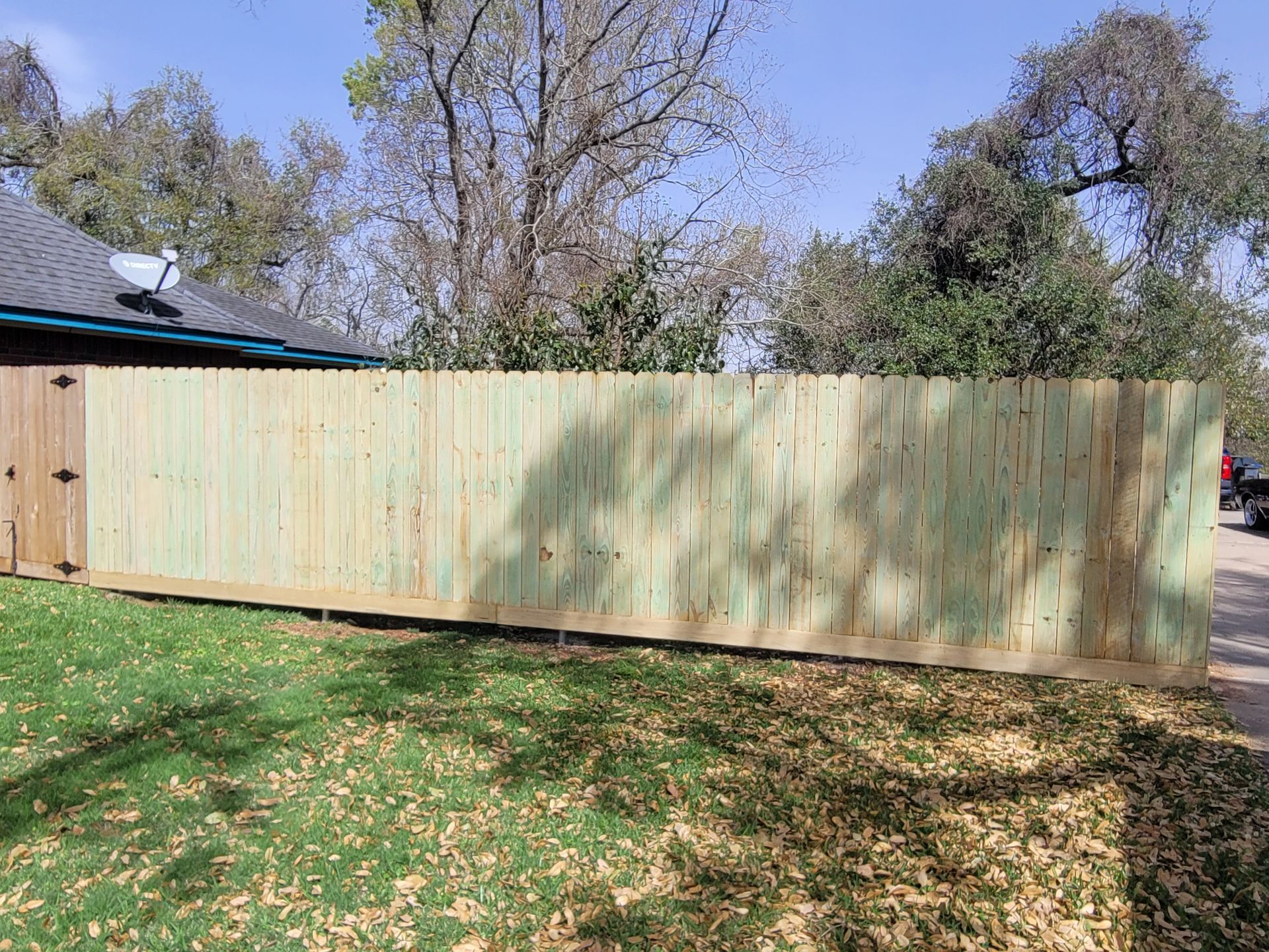 New wooden privacy fence in a backyard with trees and a house roof visible behind it.