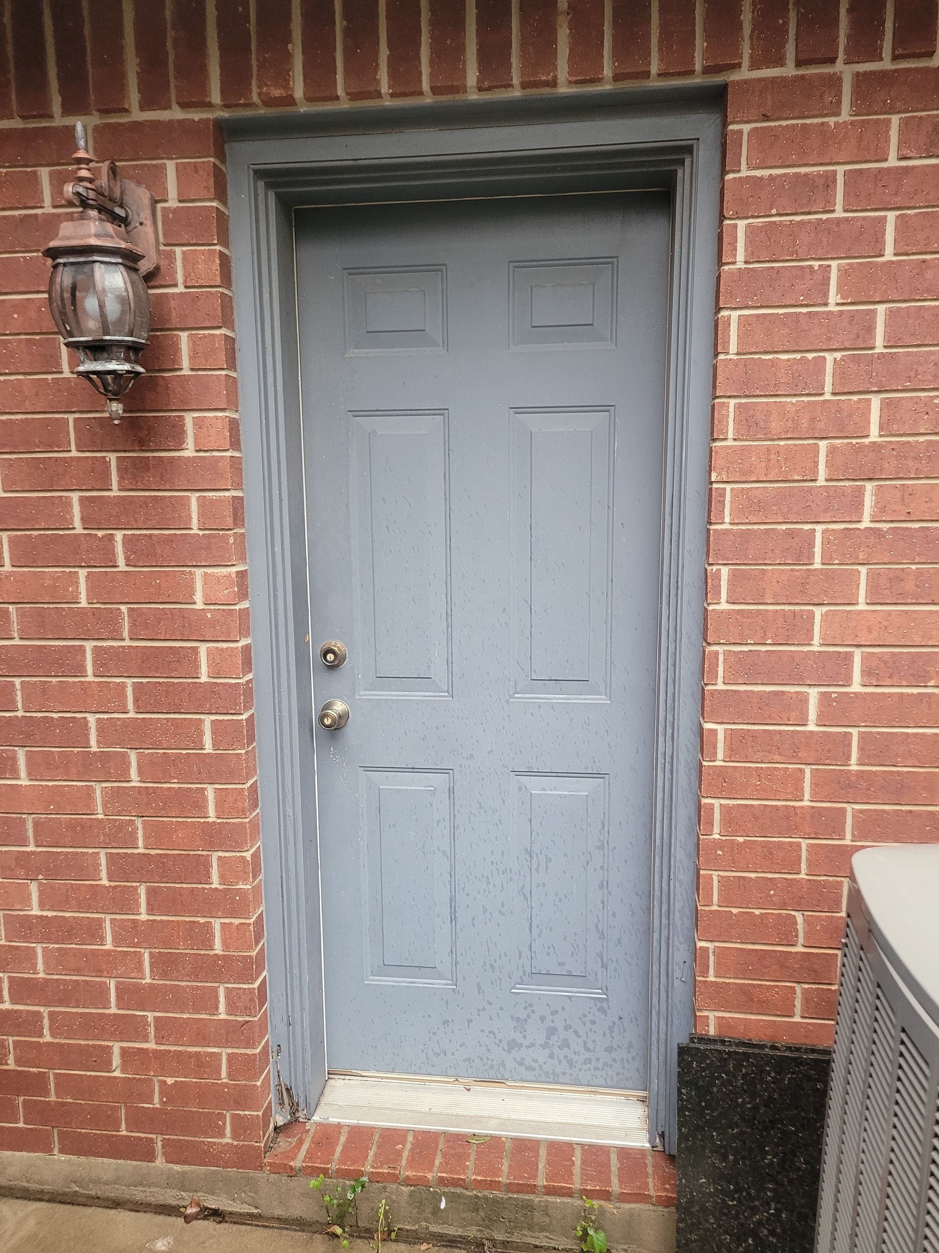 Gray front door in a red brick wall with brass knob and lantern light fixture.