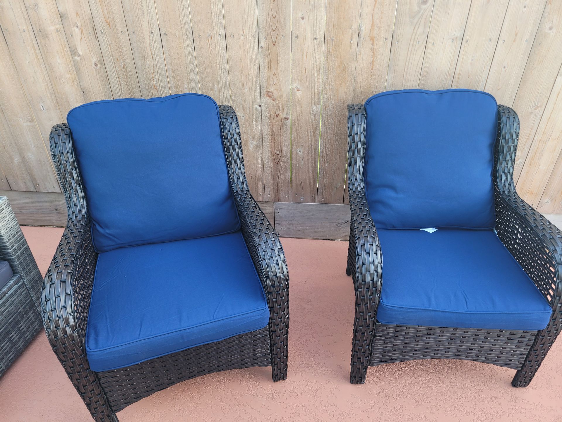 Two blue cushioned patio chairs with dark woven frames against a wooden fence.
