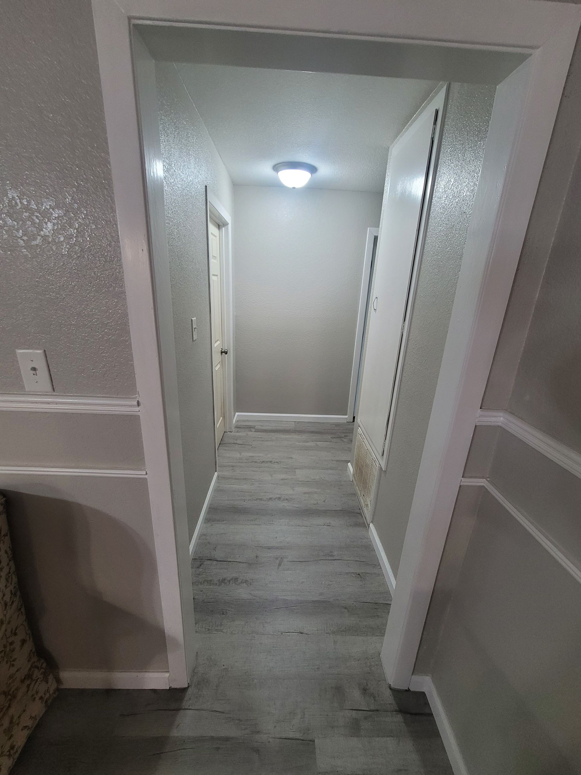Narrow hallway with gray walls, wood-look floor, and a lit ceiling lamp, viewed through a doorway