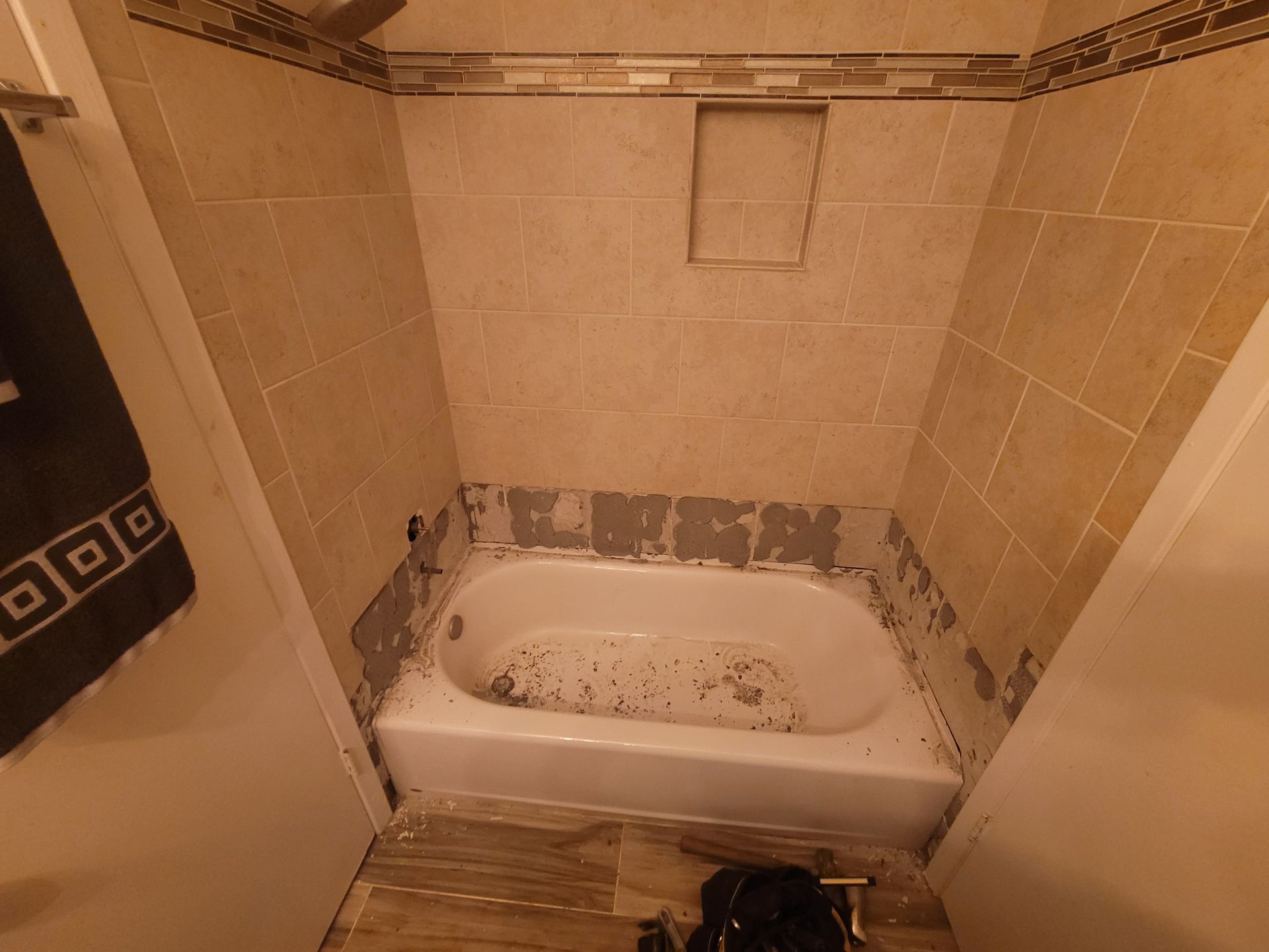 Small beige-tiled bathroom with a white bathtub and partially removed tiles around it.