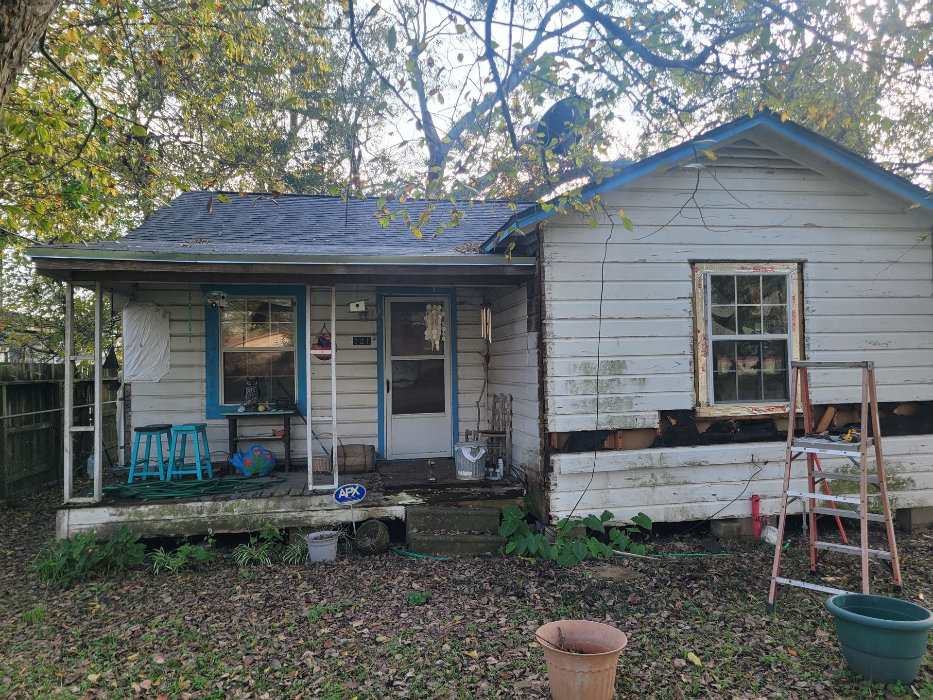 Small white house with peeling siding, porch, and scattered yard items in a wooded yard