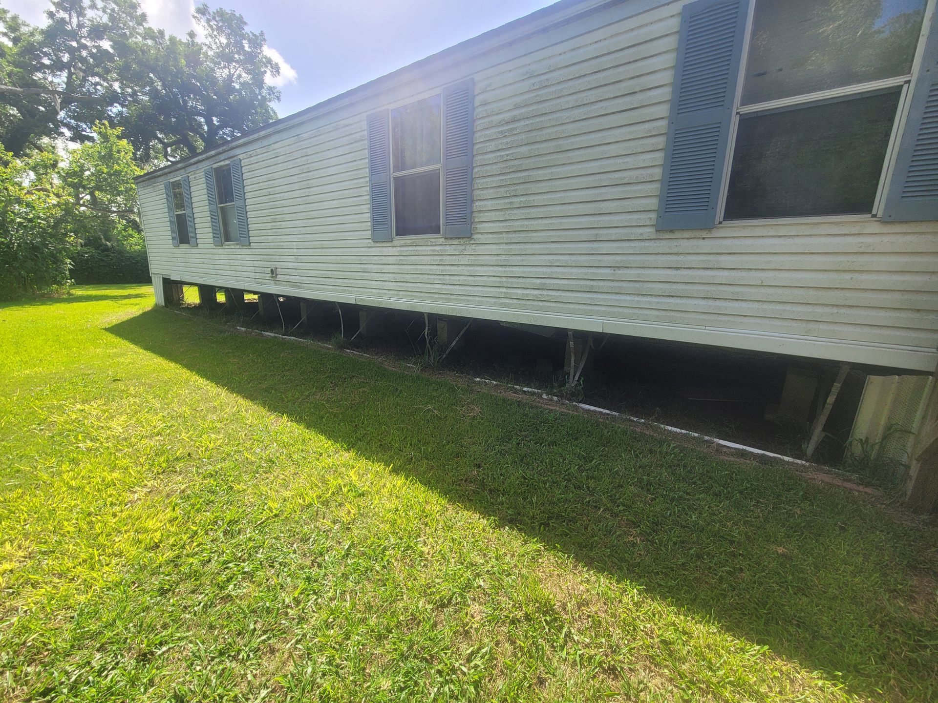 White mobile home on stilts in a grassy yard, with bright sunlight and trees in the background