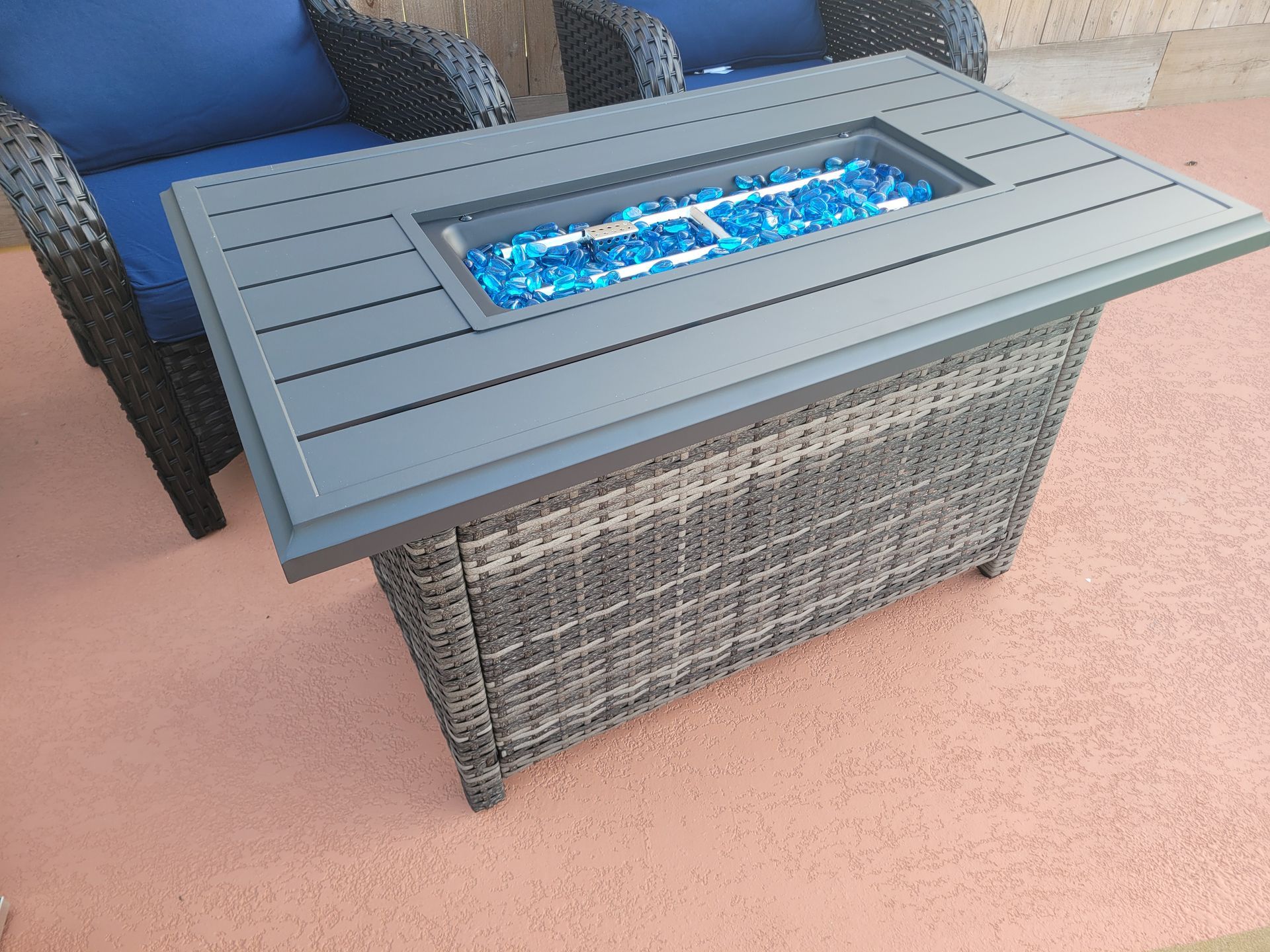 Gray wicker fire table with blue flames on a patio beside a blue chair and beige carpet