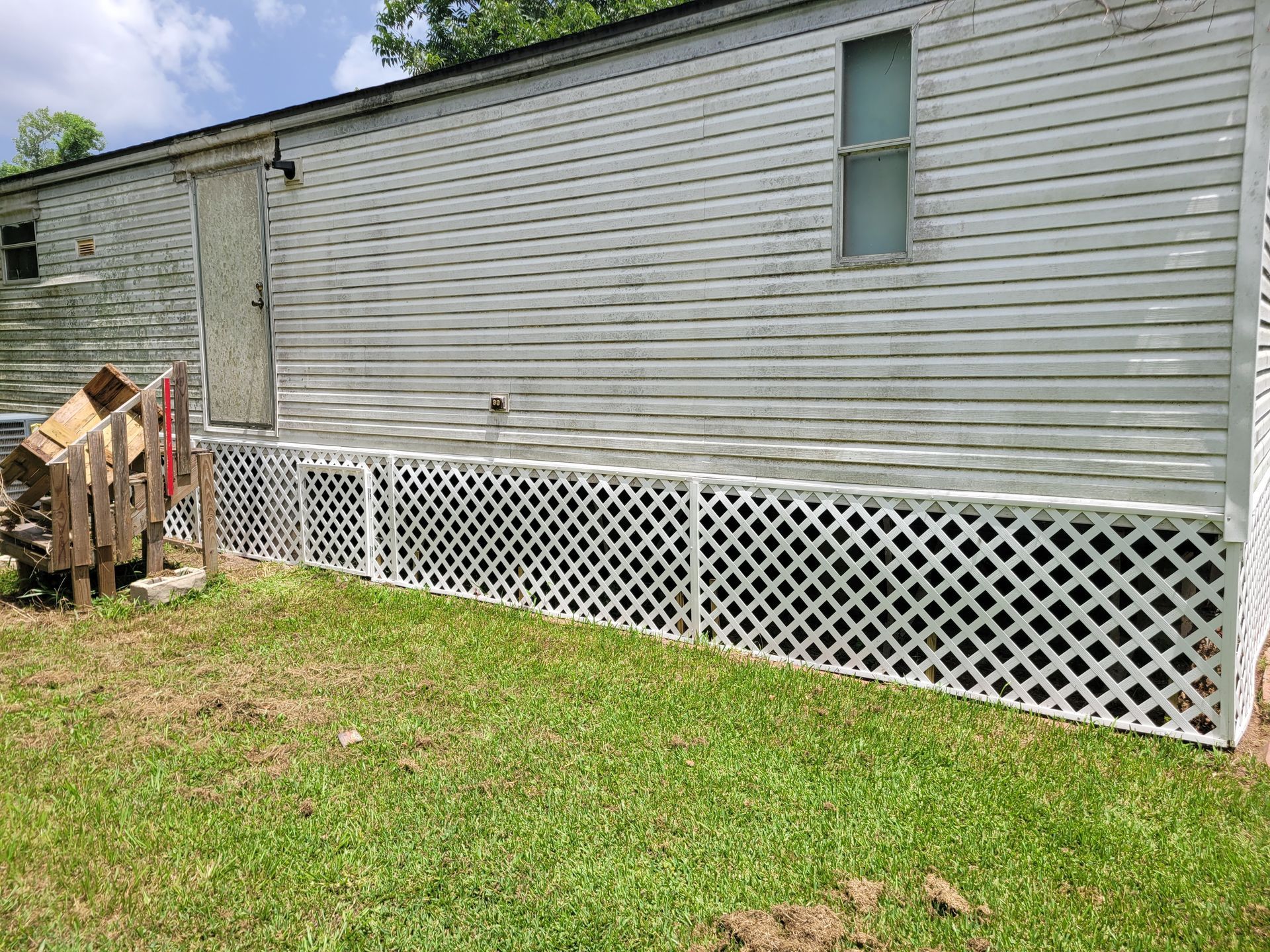 Side view of a gray mobile home with white lattice skirting and a grassy yard.