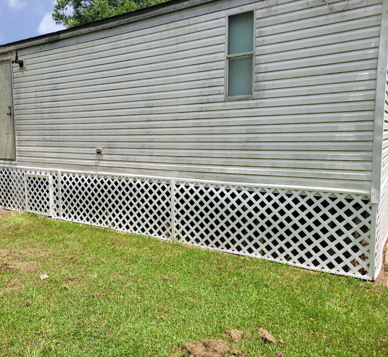 White mobile home with a small window and white lattice skirting above a grassy yard.