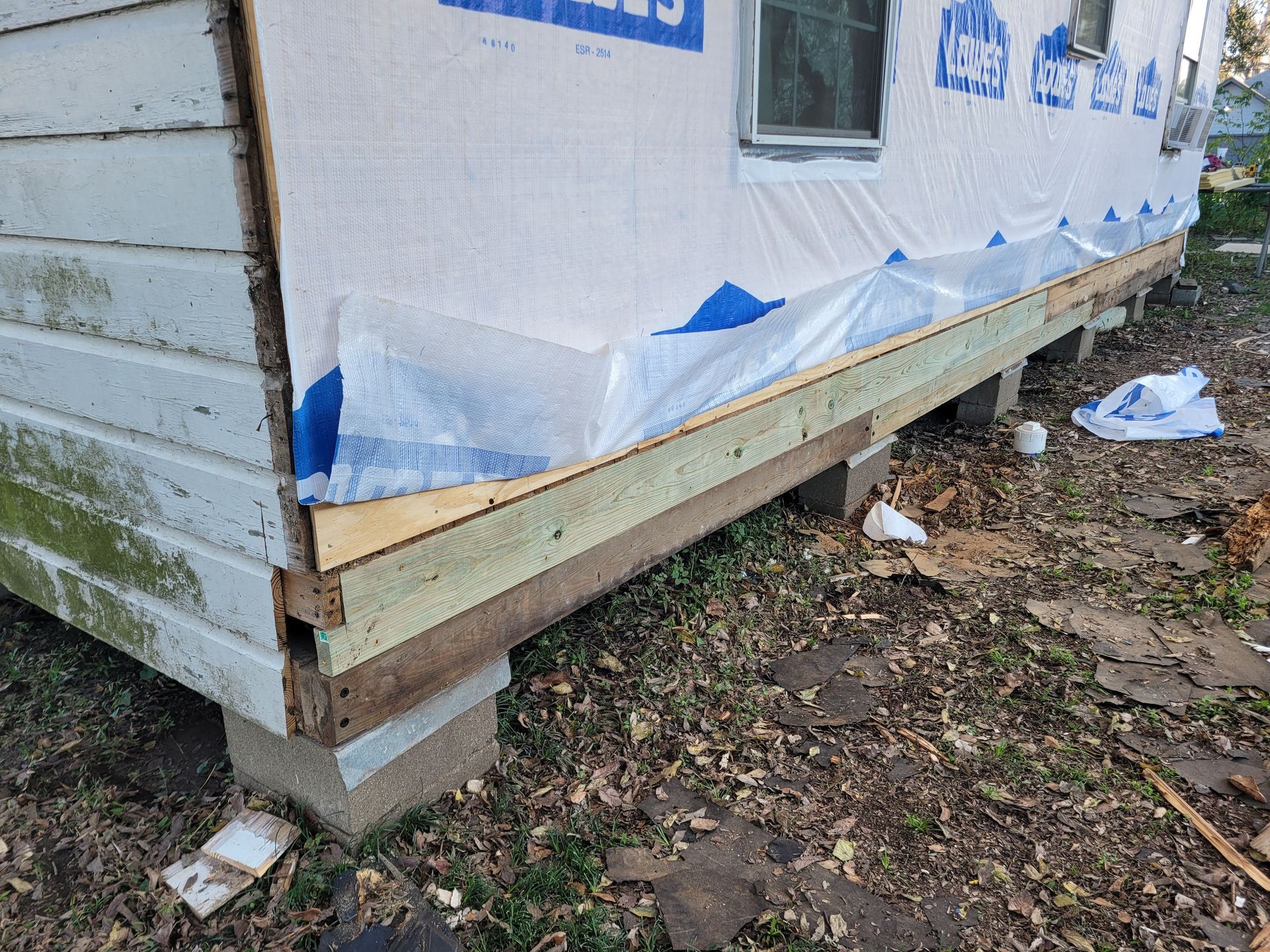 House wall under construction with house wrap, wooden trim, and concrete foundation near muddy ground