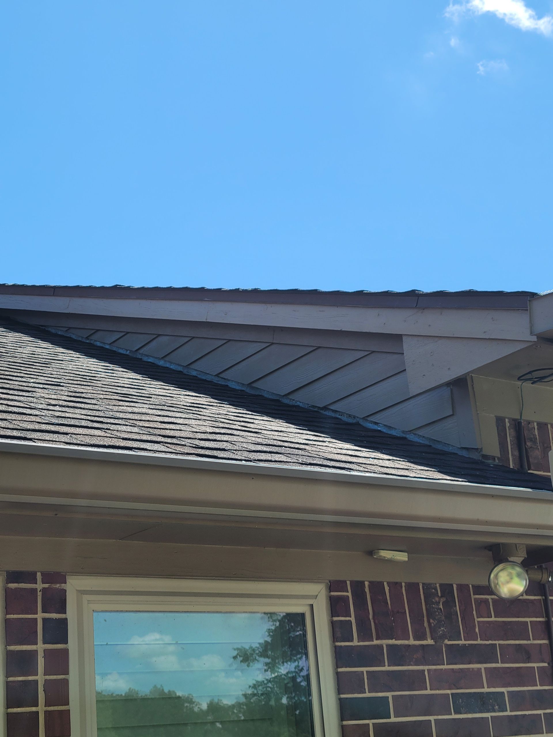 Brick house roofline with gray shingles under a bright blue sky, above a window.