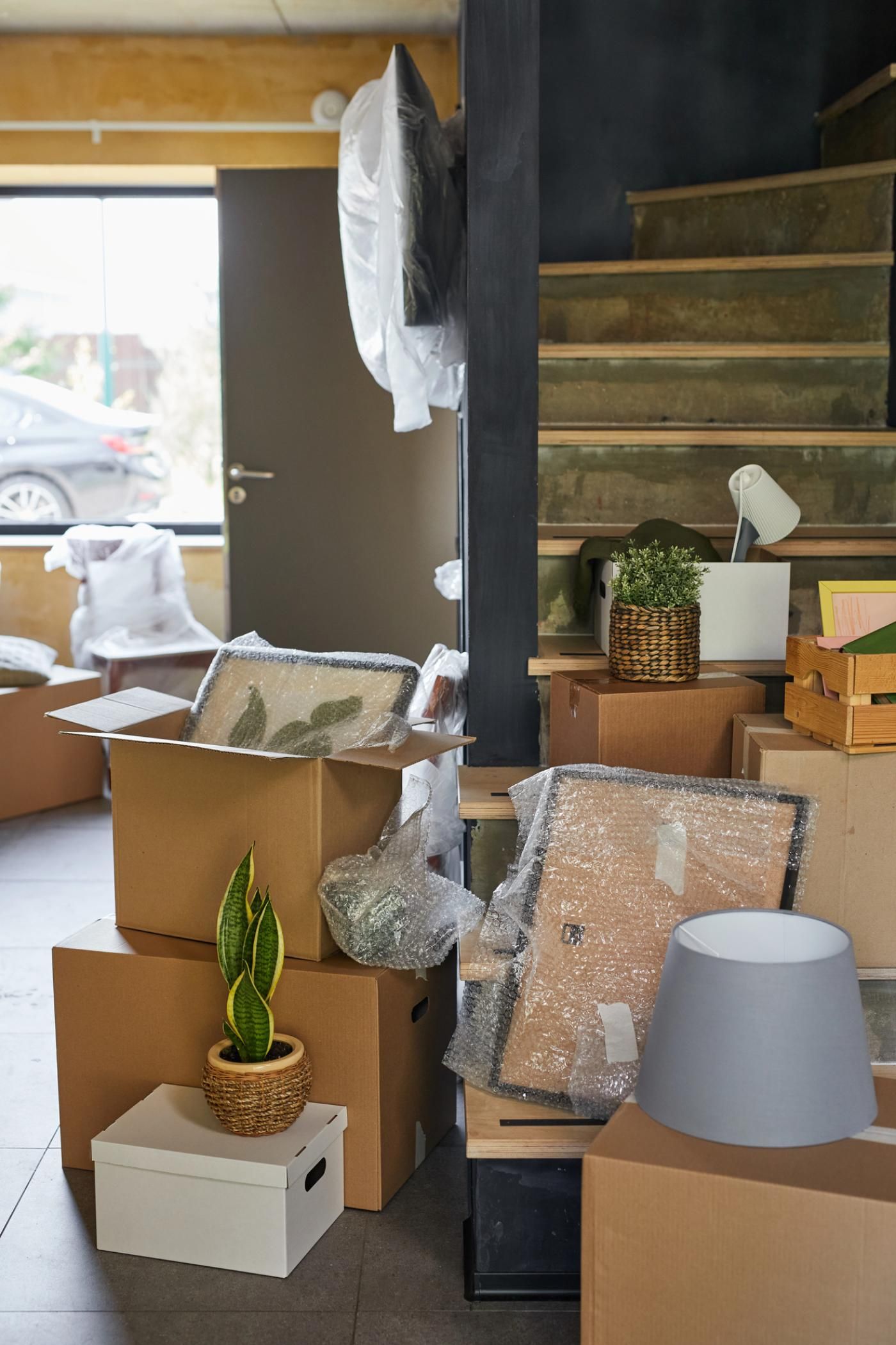 Scatole di cartone imballate per un trasloco sono accatastate vicino a una scala di legno, con piante in vaso e oggetti decorativi appoggiati sopra.