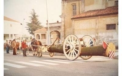 Dei cavalli trainano un carro con ruote di legno che trasporta un grosso tronco su una strada lastricata vicino a un edificio con un arco.