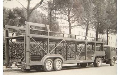 Una foto in bianco e nero di un rimorchio a due piani per il trasporto di automobili, agganciato a un camion e parcheggiato in una strada vicino ad alcuni alberi.