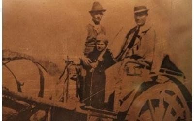 Una foto d'epoca in tonalità seppia che ritrae tre persone in posa su un carro trainato da cavalli in un ambiente rurale.