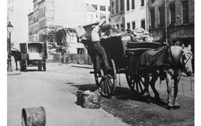Un carro trainato da cavalli viene caricato su una strada cittadina, con un'altra carrozza visibile sullo sfondo.