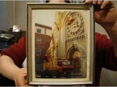 Una persona regge un quadro incorniciato raffigurante una gru gialla intenta a eseguire lavori di manutenzione sulla facciata in pietra di una chiesa.