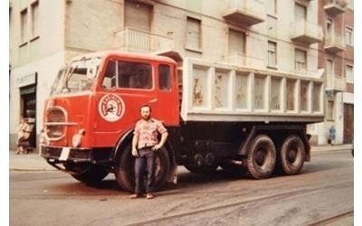 Un uomo è in piedi accanto a un grosso camion ribaltabile rosso acceso parcheggiato su una strada cittadina di fronte a un edificio.
