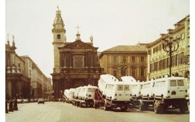 Una fila di furgoni bianchi parcheggiati in una piazza cittadina di fronte a una chiesa storica con campanile.