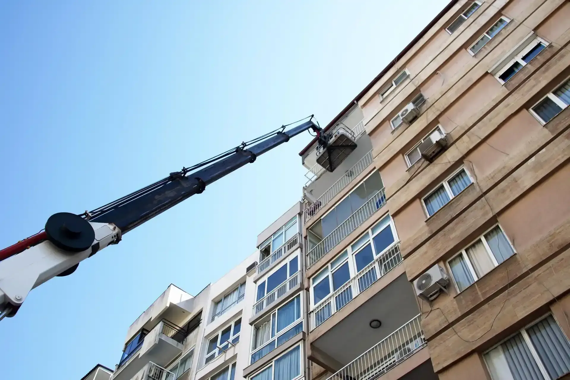 Sotto un cielo azzurro e limpido, una gru con cestello raggiunge il balcone superiore di un condominio a più piani di colore beige.