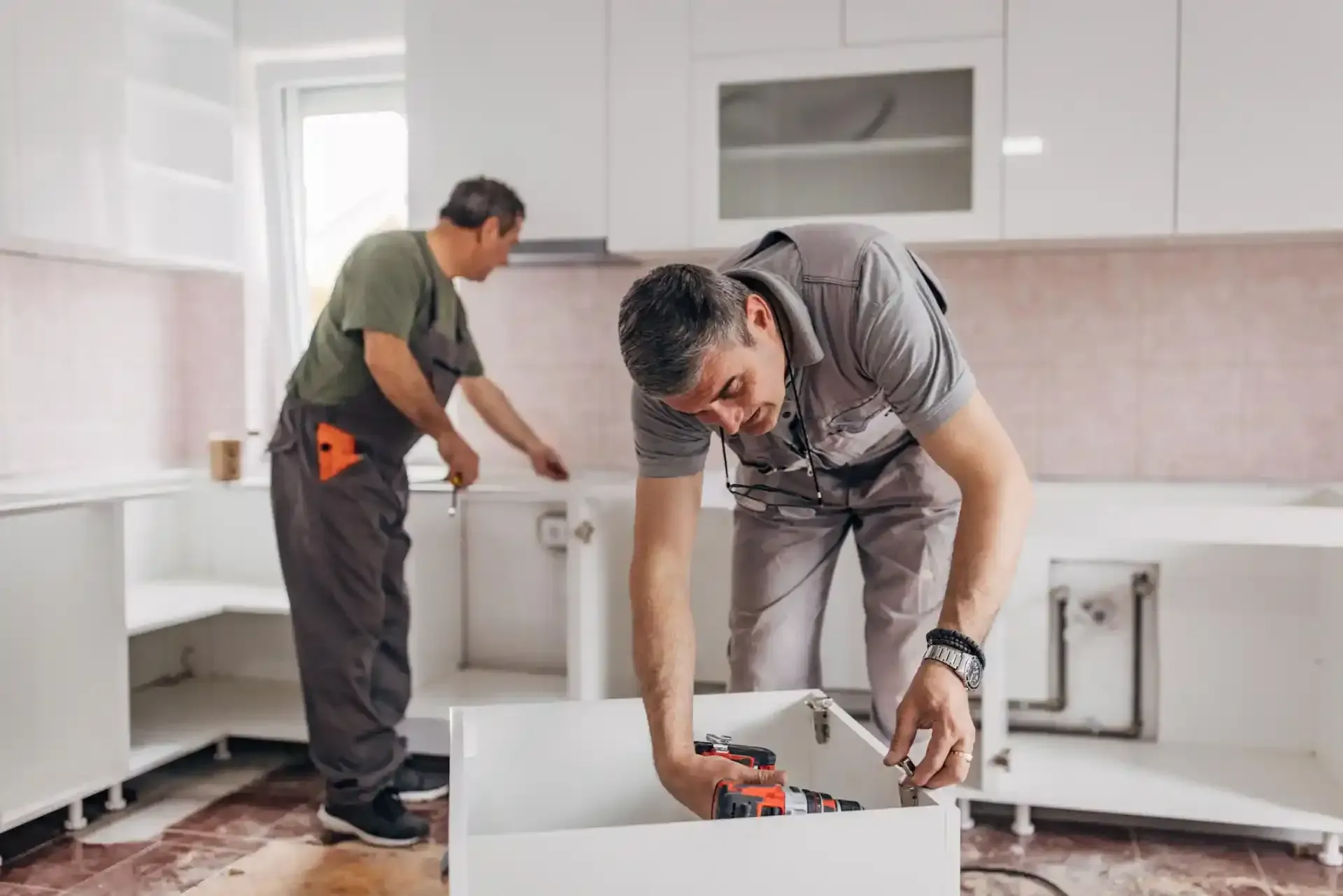 Due operai in uniforme grigia assemblano mobili da cucina bianchi in una cucina non arredata.