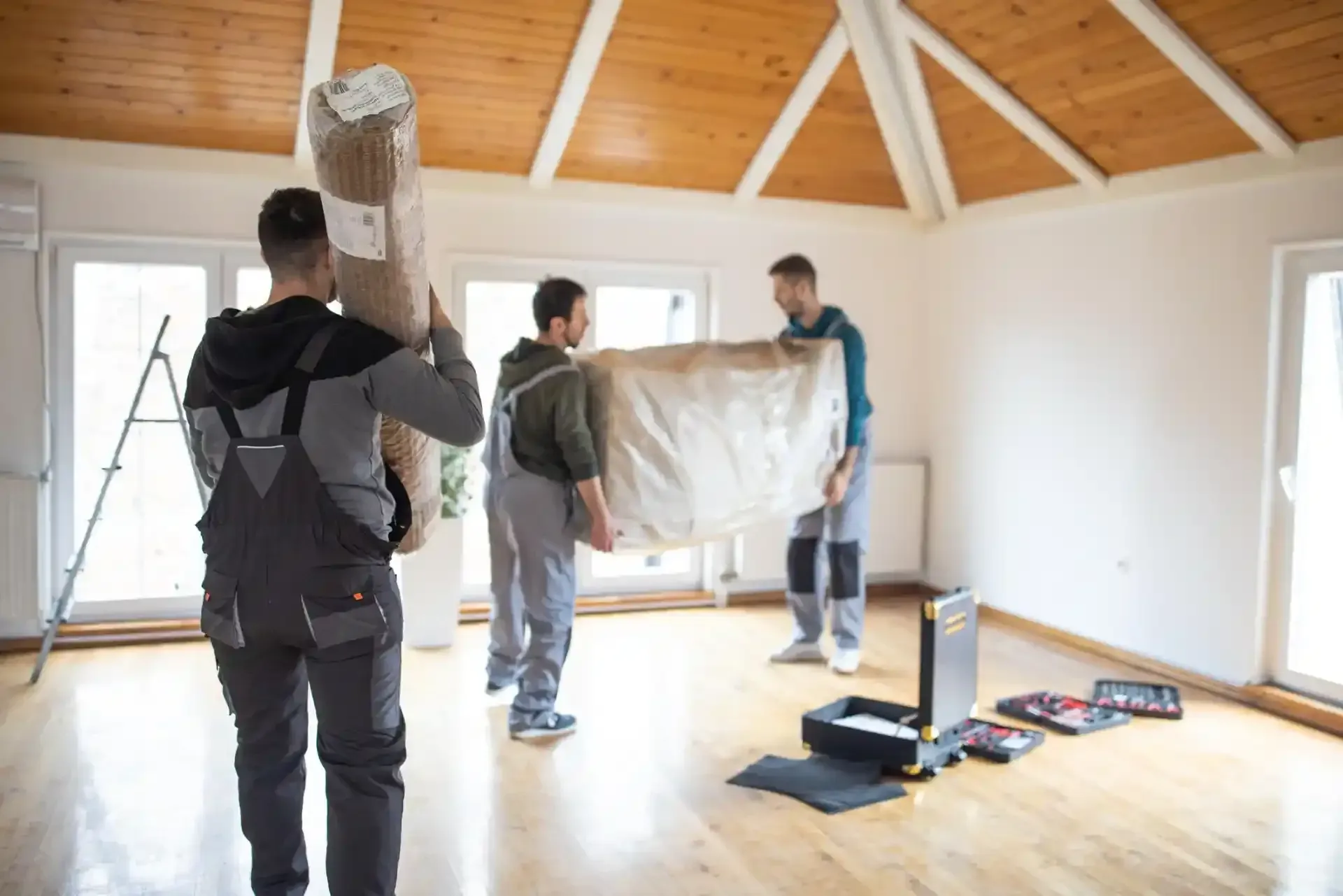 Tre traslocatori trasportano un tappeto arrotolato e un grande mobile imballato in una stanza vuota con un soffitto a volta in legno.