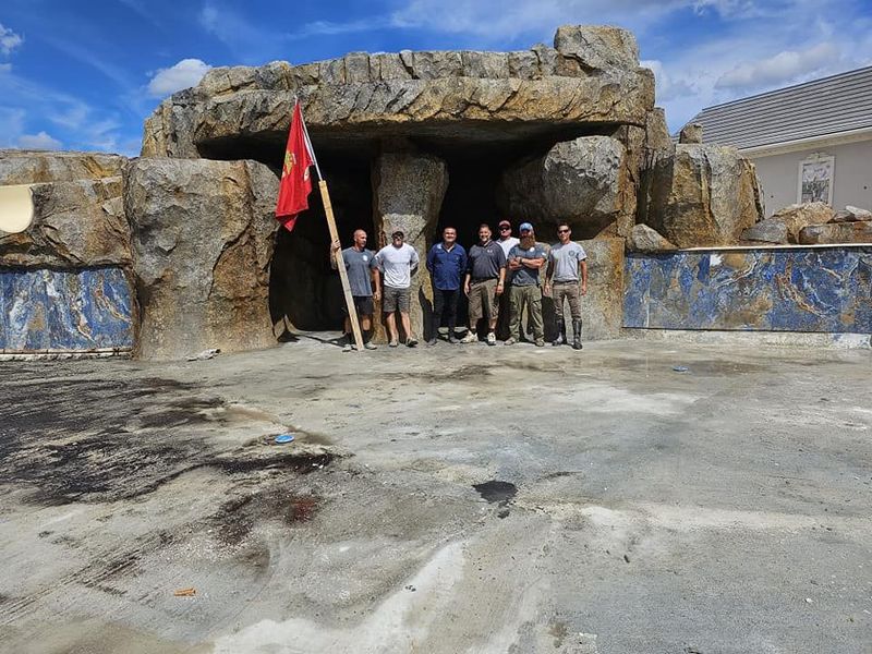 A group of men stand in front of a rock-like structure, holding a flag under a blue sky.