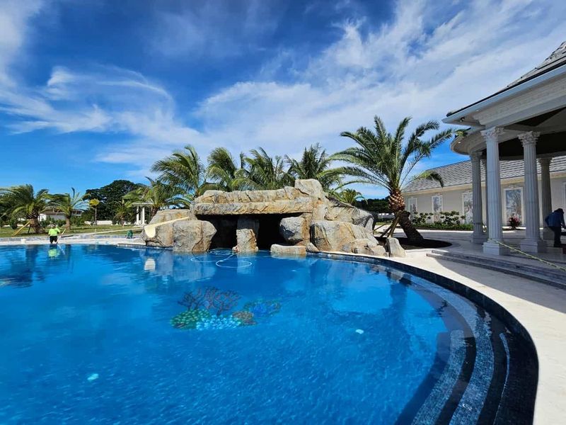 Blue swimming pool with rock waterfall feature, palm trees, and a white, columned structure. Bright blue sky.