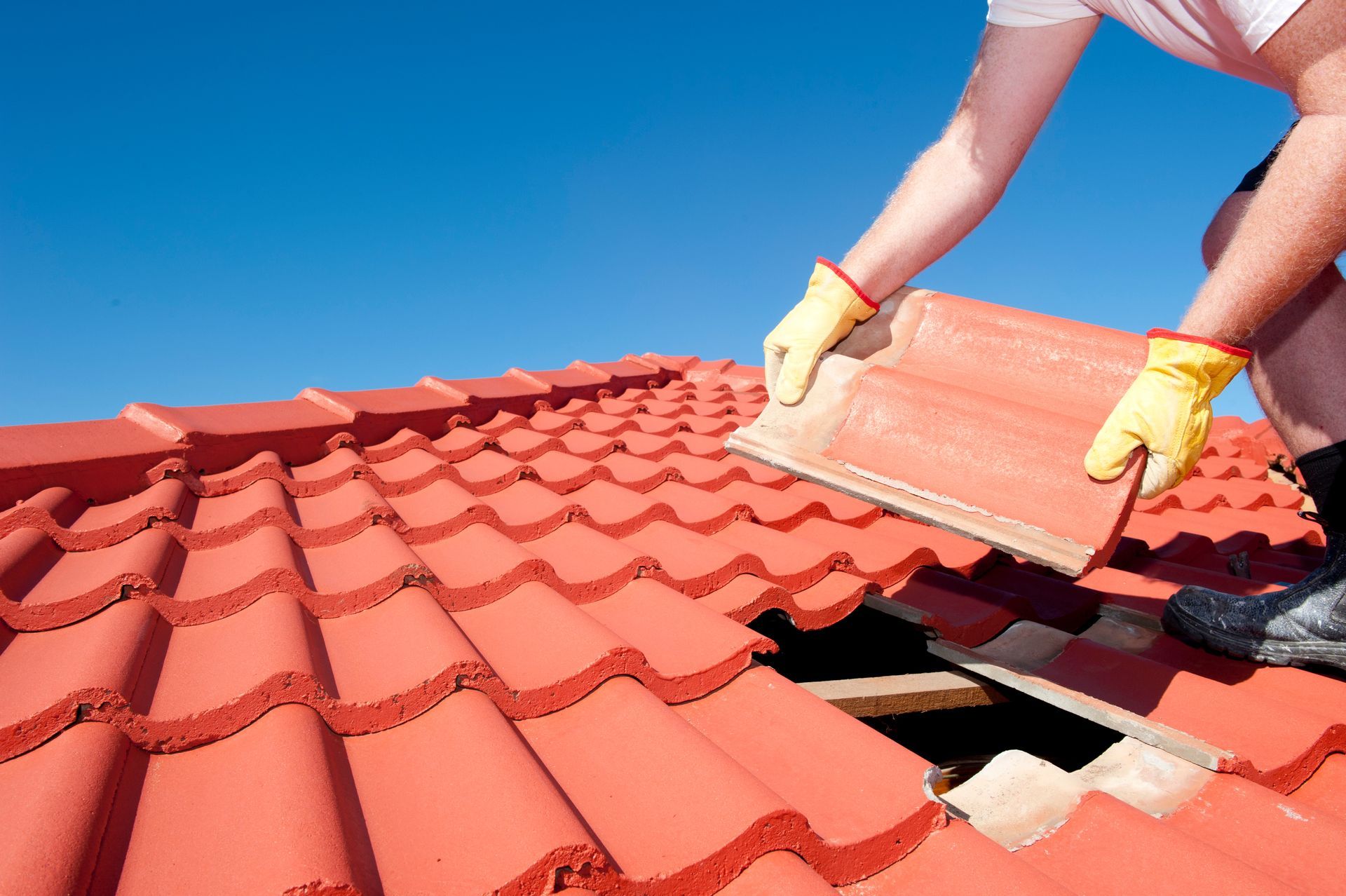 A worker wearing yellow gloves performs a residential roof repair, replacing a tile on the roof.