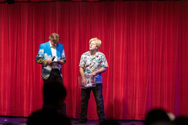 Two men are standing on a stage in front of a red curtain.