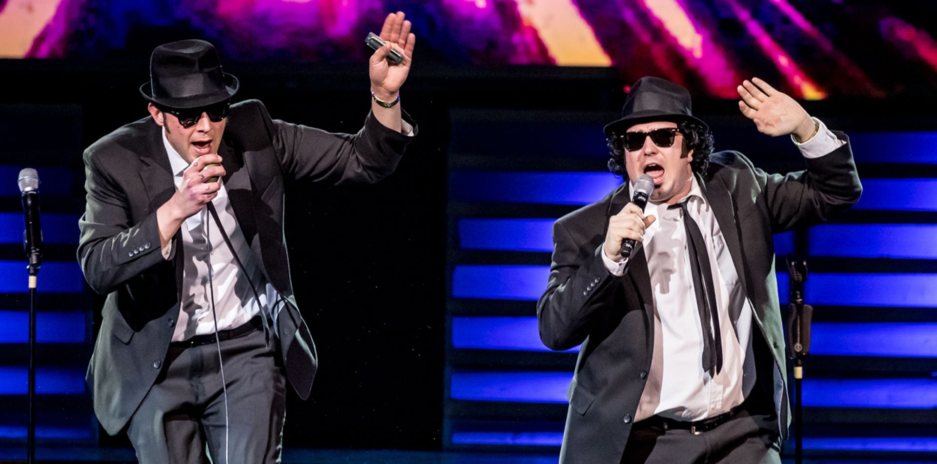 Two men in black suits, hats, and sunglasses sing into microphones on stage.