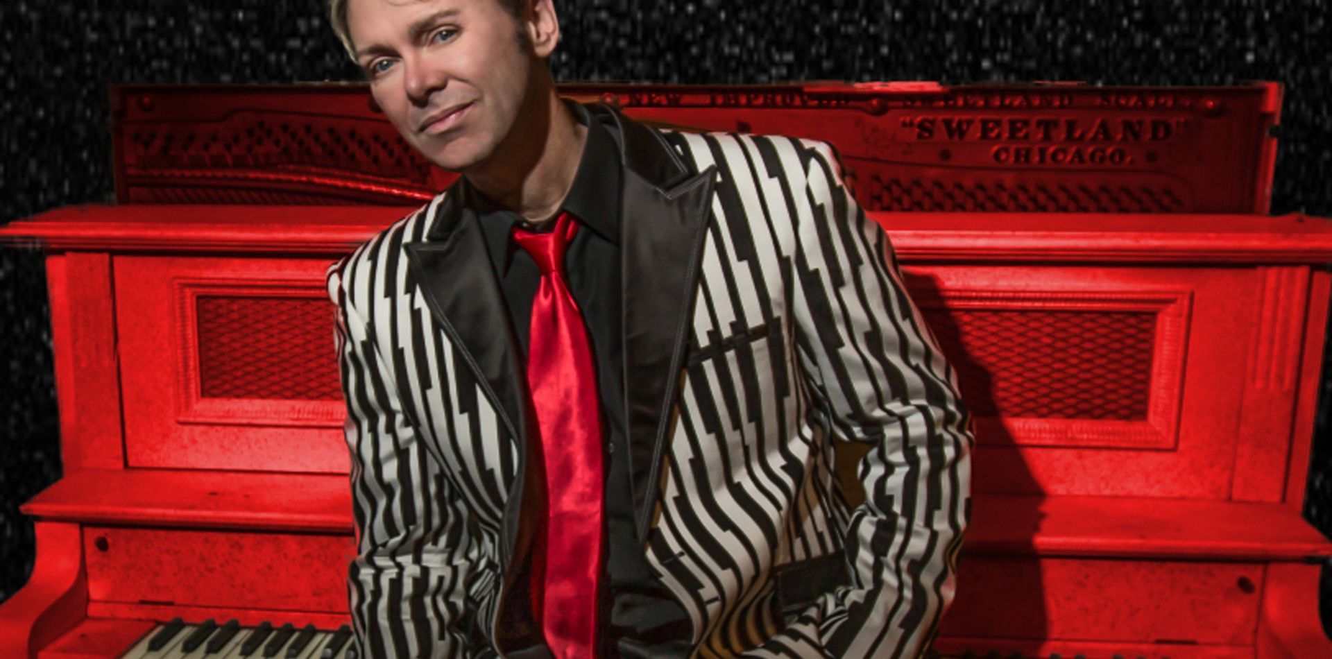 A man in a zebra print suit and tie is sitting on a red piano.