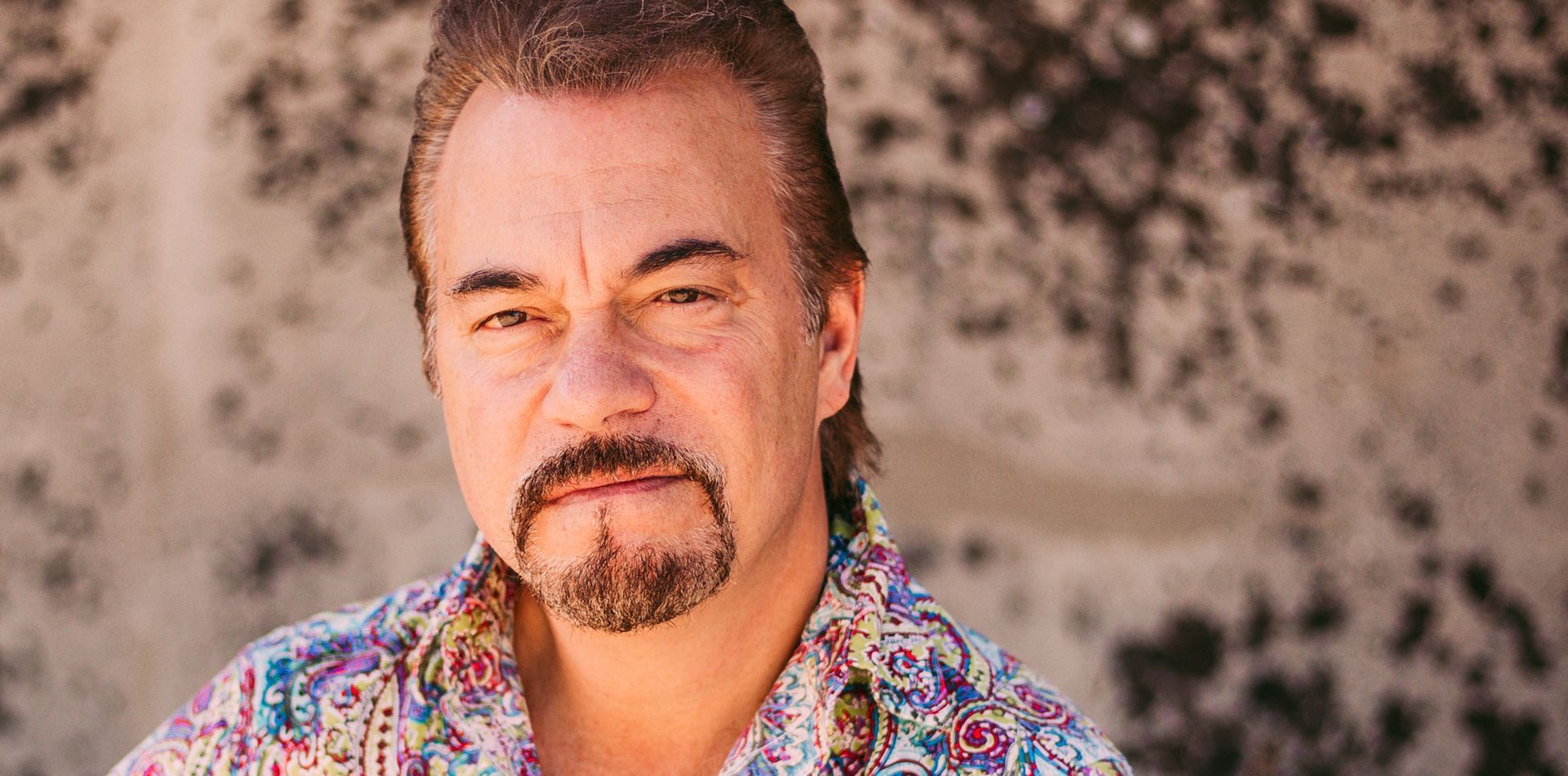 A man with a beard is wearing a colorful shirt and looking at the camera.