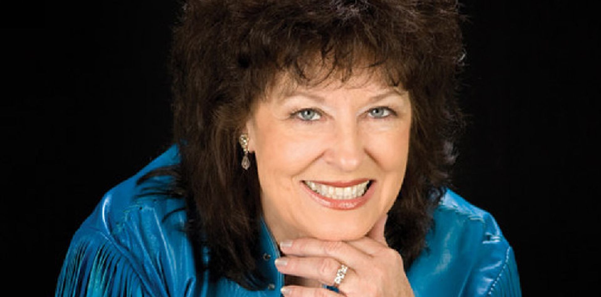 Woman with brown hair and blue top smiling, resting chin on hand. Black background.